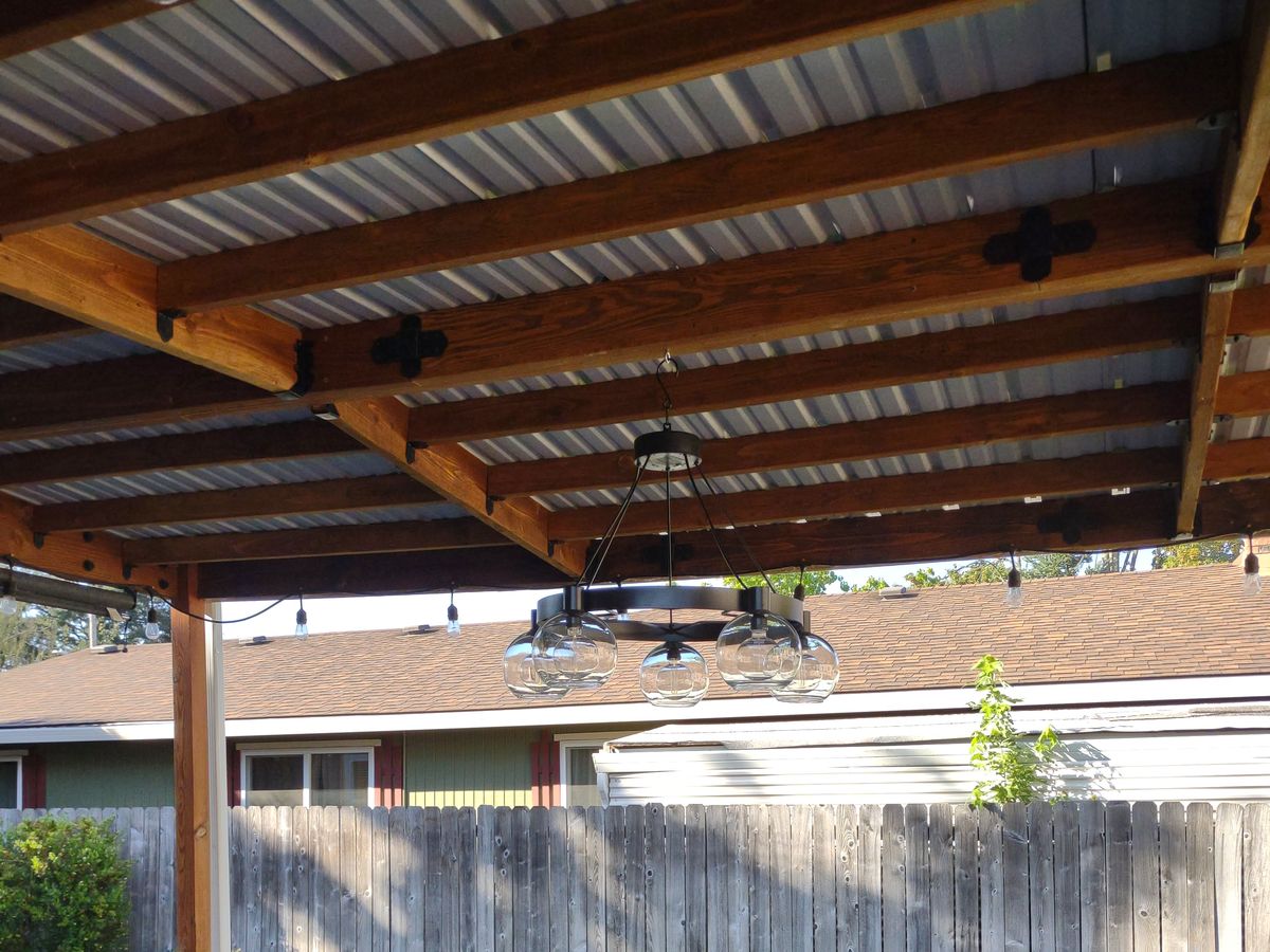Cedar-framed patio cover with corrugated metal roofing and hanging pendant lighting over a backyard patio